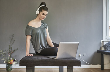 Junge Frau mit Kopfhörern, die im Wohnzimmer sitzt und einen Laptop benutzt - FMKF04015