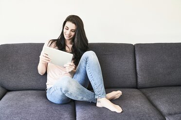 Smiling young woman sitting on couch using tablet - FMKF04042