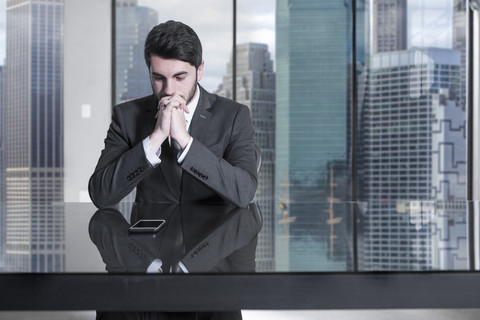 Seriöser Geschäftsmann sitzt am Schreibtisch in einem städtischen Büro und schaut auf sein Handy, lizenzfreies Stockfoto