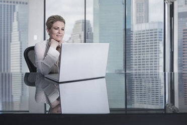 Smiling businesswoman sitting at desk in city office looking at laptop - ZEF13660