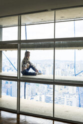 Young woman in high rise building sitting in window, looking over city - VABF01349