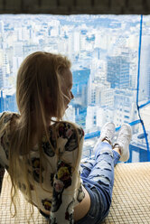 Junge Frau in einem Hochhaus, die im Fenster sitzt und über die Stadt blickt - VABF01350