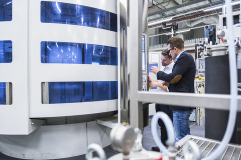 Zwei Männer unterhalten sich in einer Fabrikhalle und schauen auf einen Bildschirm, lizenzfreies Stockfoto