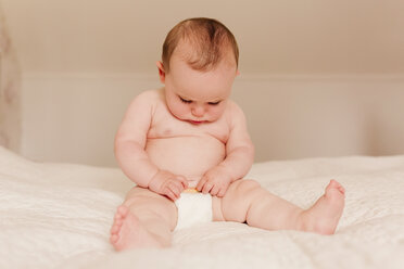 Baby girl sitting on bed examining diapers - NMSF00061