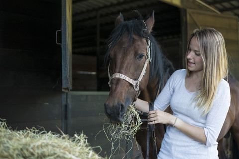 Junge Frau füttert Pferd auf dem Bauernhof mit Heu, lizenzfreies Stockfoto