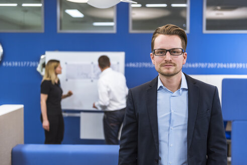 Geschäftsmann steht in einem Konferenzraum, während Kollegen im Hintergrund mit einem Flipchart arbeiten - DIGF02287