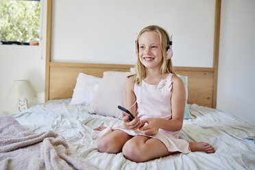 Porträt eines lächelnden kleinen Mädchens, das auf dem Bett sitzt und mit Kopfhörern Musik hört - SRYF00262