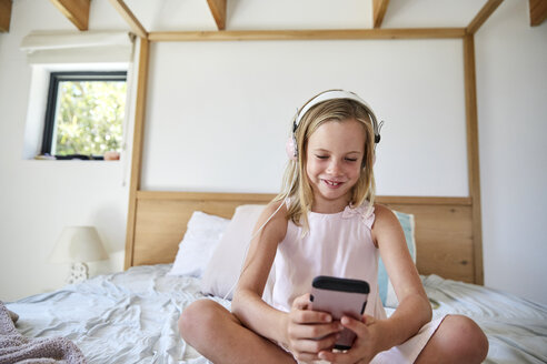 Smiling little girl sitting on bed at home while listening music with headphones - SRYF00263