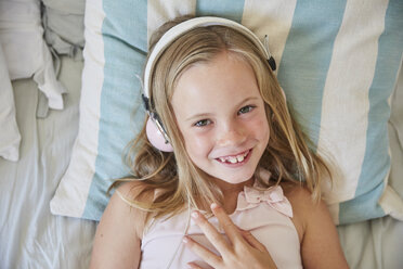 Portrait of smiling little girl lying on bed at home listening music with headphones - SRYF00265