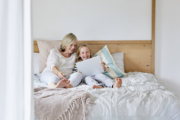 Laughing little girl lying on the bed with her grandmother using tablet - SRYF00286