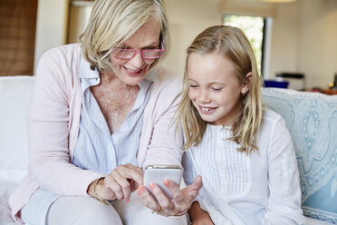 Grandmother and granddaughter sitting on the couch at home looking at smartphone - SRYF00303