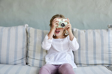 Porträt eines lächelnden kleinen Mädchens, das auf der Couch sitzt und ein Foto mit der Kamera macht - SRYF00324