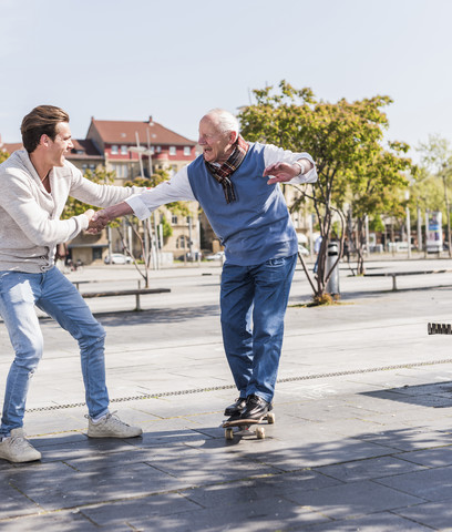 Erwachsener Enkel unterstützt älteren Mann auf Skateboard, lizenzfreies Stockfoto