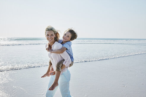 Happy mother carrying son piggyback on the beach - RORF00787