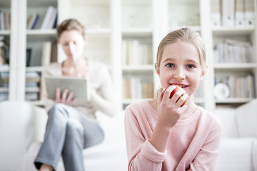 Girl eating apple at home with mother in background - WESTF23039