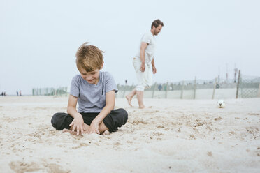 Lächelnder kleiner Junge, der auf dem Sand am Strand sitzt, während sein Vater im Hintergrund mit einem Ball spielt - MVCF00157