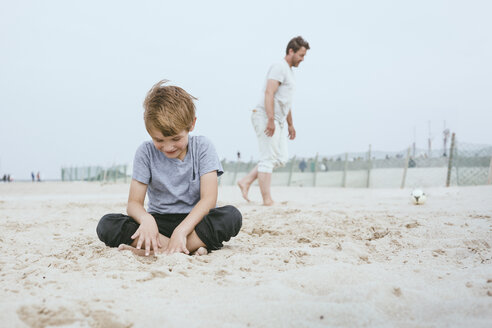 Smiling little boy sitting on the sand on the beach while his father playing with ball in the background - MVCF00157