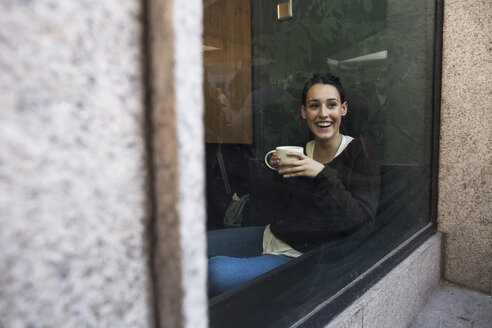 Porträt einer lächelnden Frau mit einer Tasse Kaffee, die hinter einer Fensterscheibe in einem Café sitzt - ABZF01971