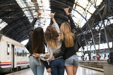 Rückenansicht von drei jungen Frauen, die Arm in Arm auf einem Bahnsteig stehen - KKAF00724