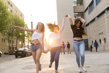 Three happy young women running hand in hand on sidewalk - KKAF00737