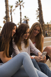 Drei lächelnde junge Frauen sitzen auf einer Treppe und schauen auf ihr Handy - KKAF00739