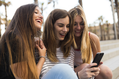 Three happy young women sitting on stairs having fun with cell phone stock photo