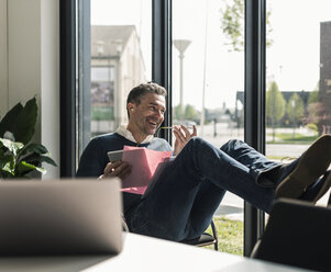 Smiling businessman sitting with feet up in his office using smartphone and earphones - UUF10494