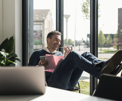 Lächelnder Geschäftsmann, der mit hochgelegten Füßen in seinem Büro sitzt und Smartphone und Kopfhörer benutzt - UUF10494