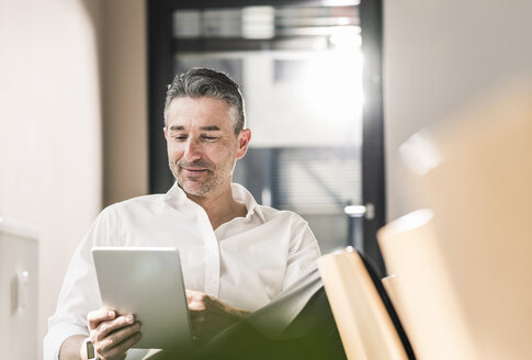 Zufriedener Geschäftsmann, der in seinem Büro sitzt und ein Tablet benutzt - UUF10501