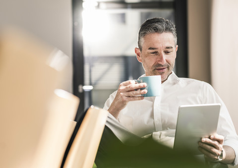 Porträt eines Geschäftsmannes mit einer Tasse Kaffee, der in seinem Büro sitzt und ein Tablet benutzt, lizenzfreies Stockfoto