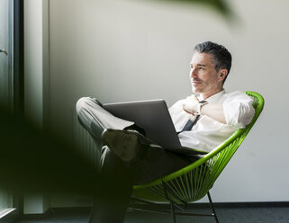 Smiling businessman with laptop sitting in an armchair - UUF10510
