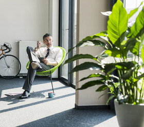 Businessman with tablet sitting in an armchair using smartphone and earphone for calling - UUF10512