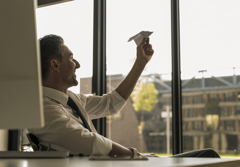 Businessman sitting at desk in his office looking at paper plane - UUF10527