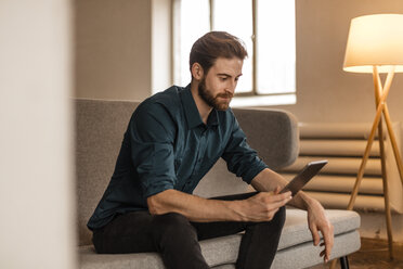 Junger Mann sitzt auf der Couch und benutzt ein Tablet - JOSF00728