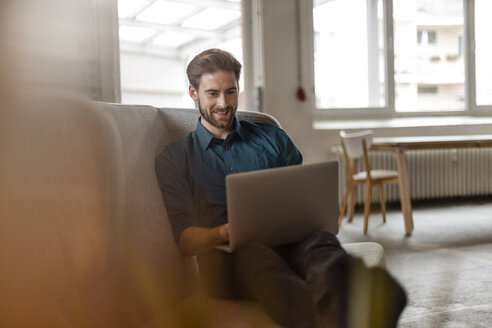 Porträt eines jungen Freiberuflers, der auf einer Couch in einem Loft sitzt und einen Laptop benutzt - JOSF00729