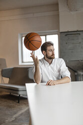 Portrait of pensive young businessman sitting at table in a loft balancing basketball on his finger - JOSF00731