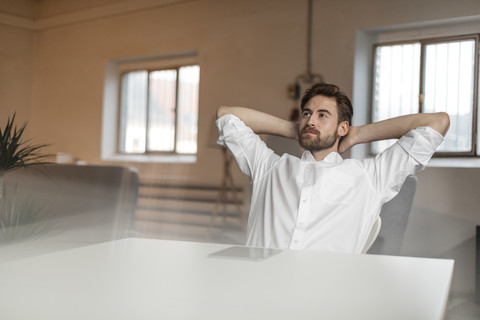 Porträt eines nachdenklichen jungen Mannes mit den Händen hinter dem Kopf am Tisch sitzend, lizenzfreies Stockfoto