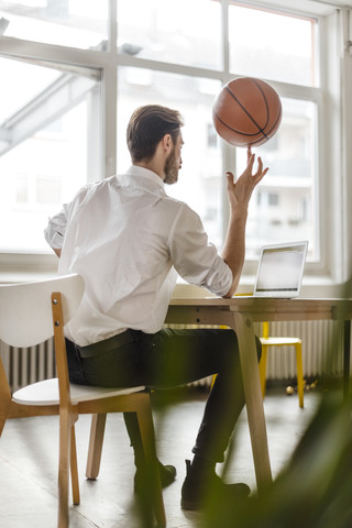Rückenansicht eines jungen Mannes, der am Schreibtisch sitzt und auf einen Laptop schaut, während er einen Basketball auf seinem Finger balanciert, lizenzfreies Stockfoto