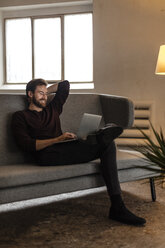 Lachender junger Mann sitzt auf der Couch und schaut auf den Laptop - JOSF00749