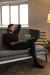 Laughing young man sitting on couch looking at laptop - JOSF00749
