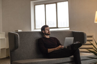 Young man sitting with laptop on couch looking at distance - JOSF00750