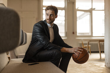 Portrait of young businessman with basketball sitting on the couch in a loft - JOSF00763