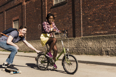 Young woman on bicycle pulling young man, standing on skateboard - UUF10556