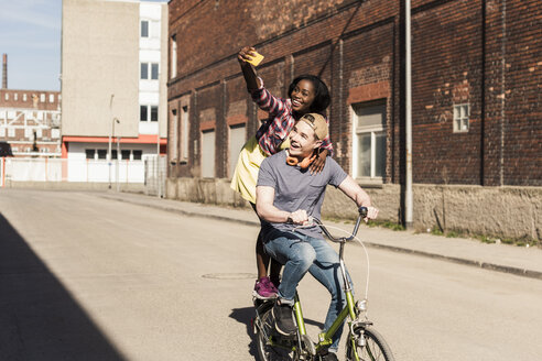 Young couple riding bicycle in the street, woman standing on rack, taking selfies - UUF10562