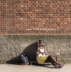 Young woman sitting on ground, listening music with headphones - UUF10569