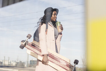 Young woman carrying skateboard, drining from disposable cup - UUF10574