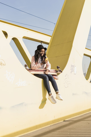 Junge Frau sitzt auf einer Brücke und benutzt ein Smartphone, lizenzfreies Stockfoto