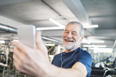 Glücklicher älterer Mann mit Smartphone und Kopfhörern im Fitnessstudio - HAPF01651