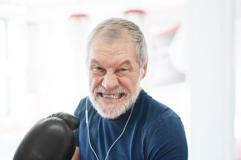 Portrait of aggressive senior man with earphones and boxing gloves in gym - HAPF01671