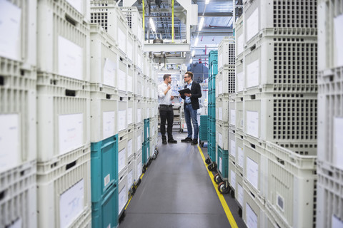 Zwei Männer mit Tablet im Gespräch über Kisten in einer Fabrikhalle, lizenzfreies Stockfoto